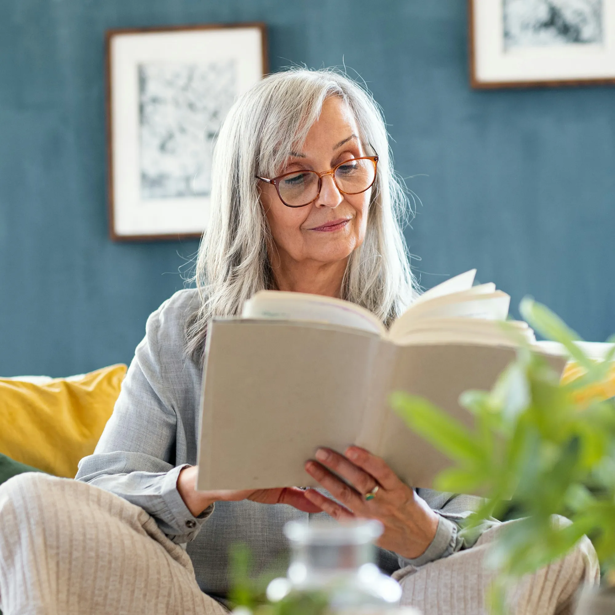Older woman with gray hair and glasses reading a book while seated on a couch.