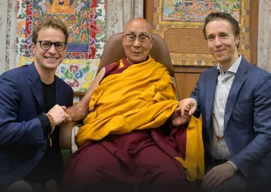 The Dalai Lama seated between two men in suits, holding hands with both.