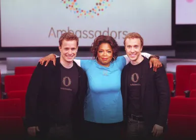 Three people standing with arms around each other in front of a screen displaying the word 'Ambassadors'.