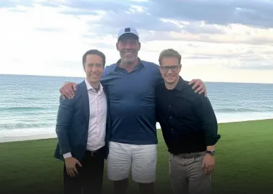 Three men standing arm-in-arm on grass by the ocean, smiling under a cloudy sky.