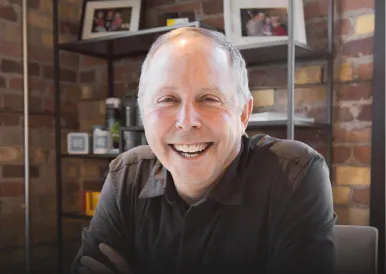 Smiling middle-aged man with short gray hair sitting indoors with brick wall and framed photos in the background.