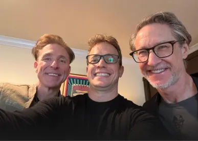 Three middle-aged men smiling for a selfie indoors with a light-colored wall and framed artwork in the background.