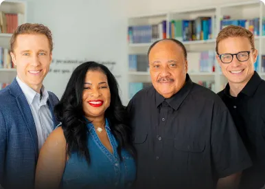 Four people smiling and standing together in a bright room with bookshelves in the background.