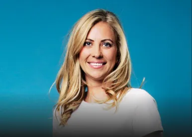 Smiling blonde woman with wavy hair wearing a white top against a blue background.