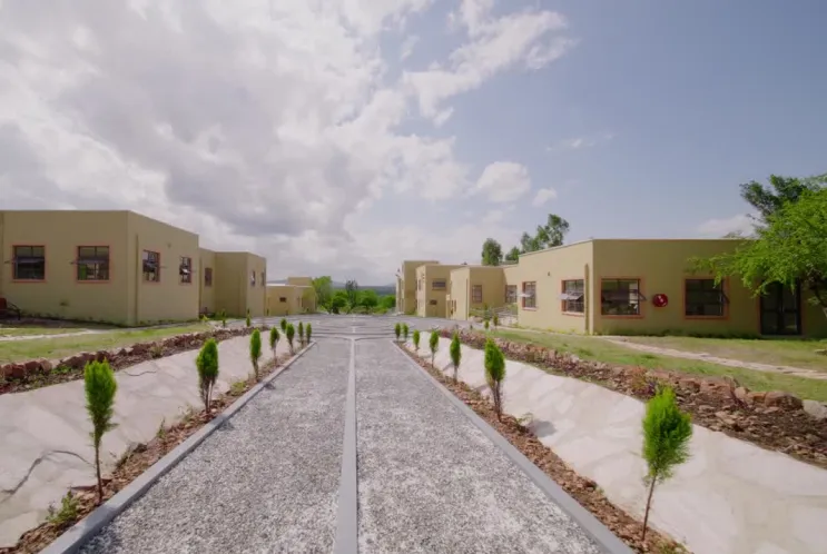 Gravel walkway lined with small trees leading through a residential area with single-story beige buildings under a partly cloudy sky.