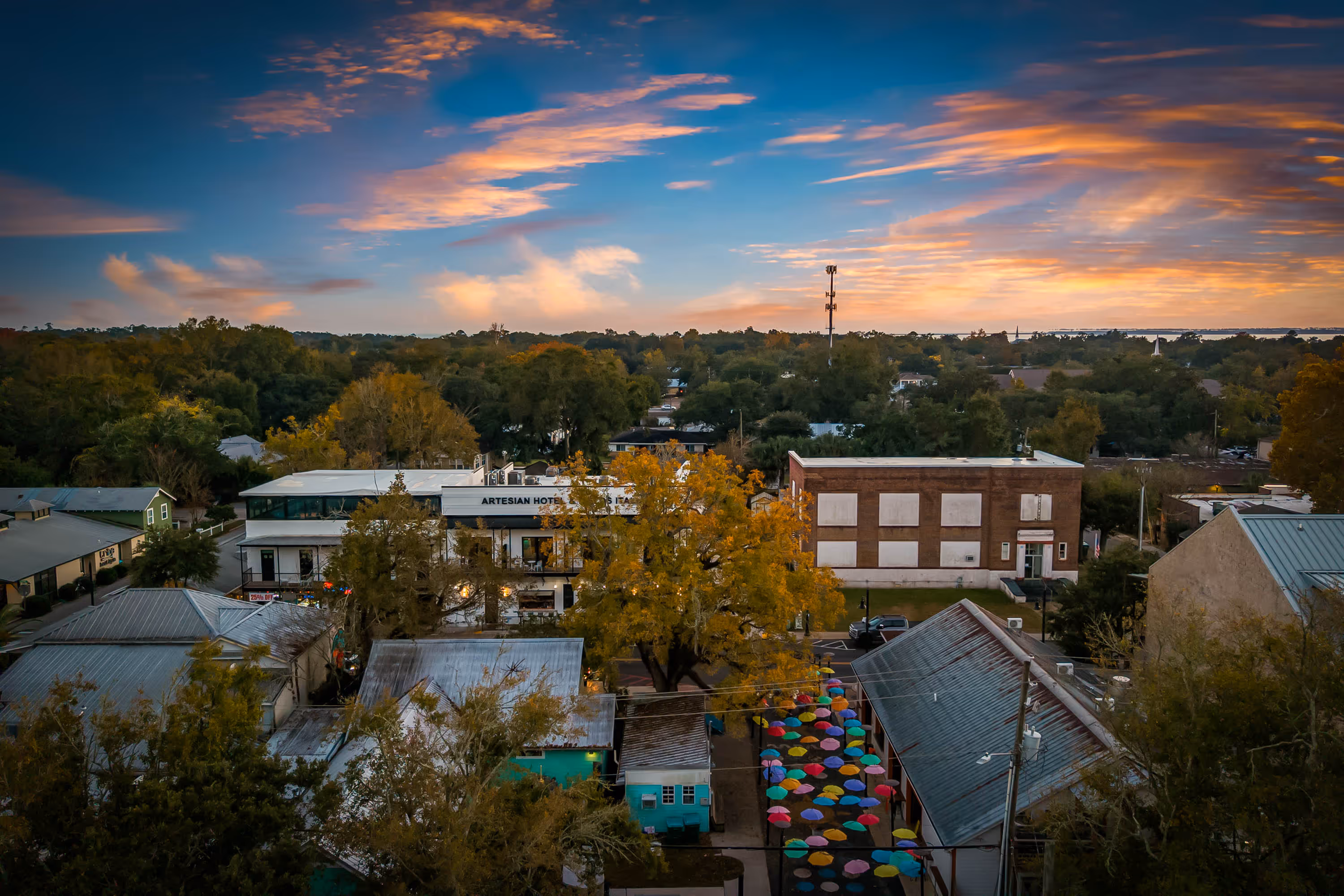 Aerial view of a small town at sunset with trees, buildings including Artesian Hotel, and a street decorated with colorful umbrellas.