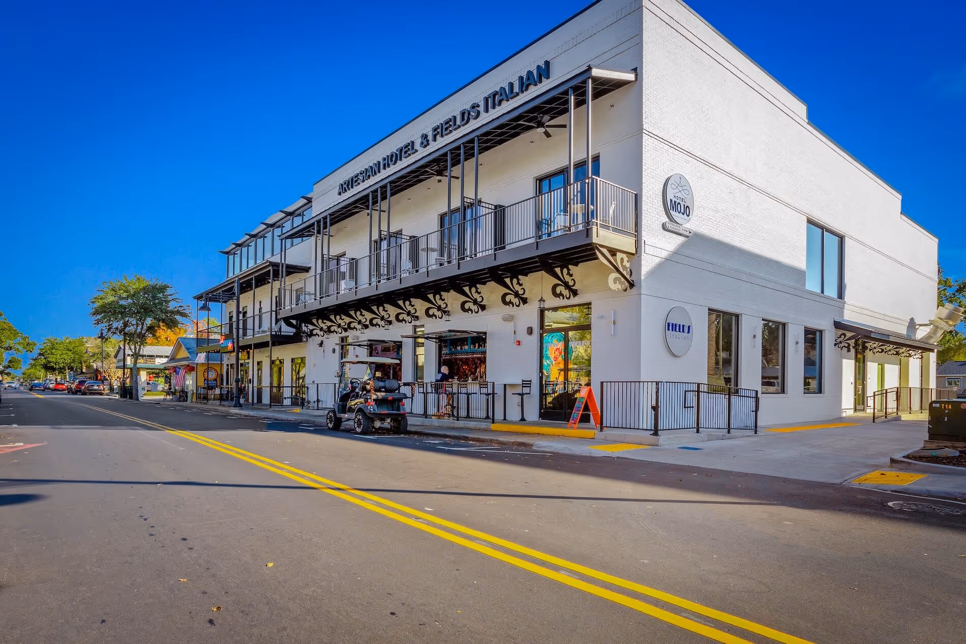 White two-story building housing Artesian Hotel and Fields Italian restaurant on a sunny street with clear blue sky.