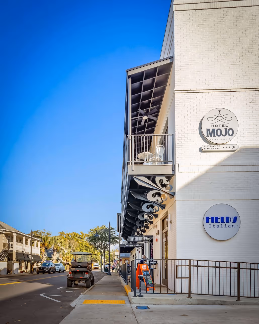 Street view of a building with white brick exterior featuring signs for Hotel Mojo and Field's Italian restaurant, a balcony with chairs, and a golf cart parked on the road.
