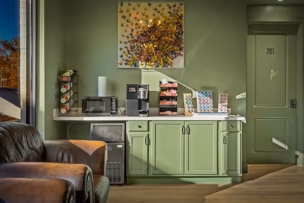Cozy coffee bar setup with a coffee machine, microwave, cups, and pods on a green cabinet, next to brown leather chairs and a door numbered 201.