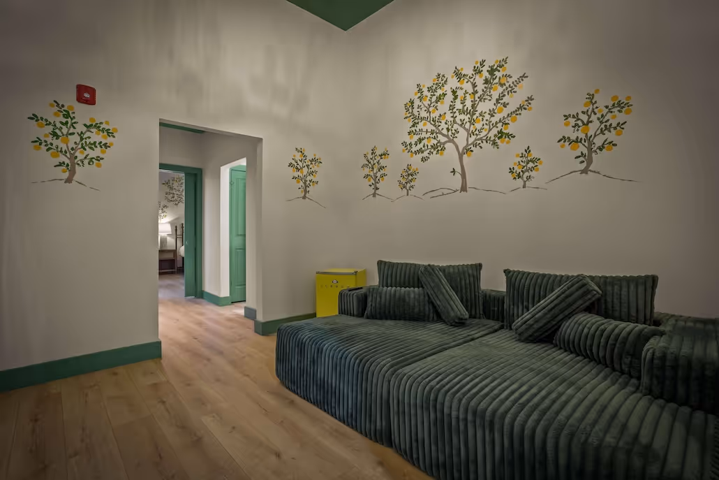 Living room with dark green ribbed sofa, yellow mini fridge, light wood floor, and walls painted with yellow-fruited trees.
