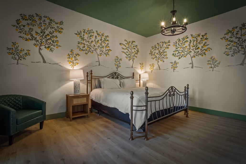 Bedroom with a wrought iron bed, white bedding, green armchair, wooden nightstand with lamp, and walls painted with a mural of lemon trees.