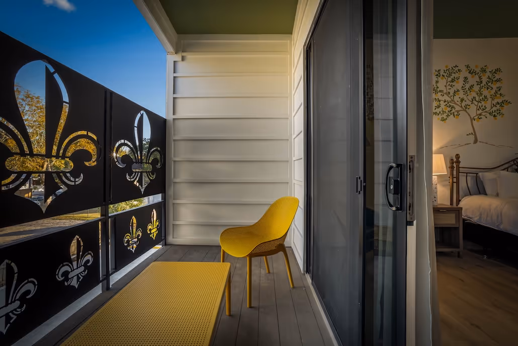 Balcony with yellow chair and table, decorative black railing with fleur-de-lis cutouts, and view into a bedroom with wall art and a lit lamp.