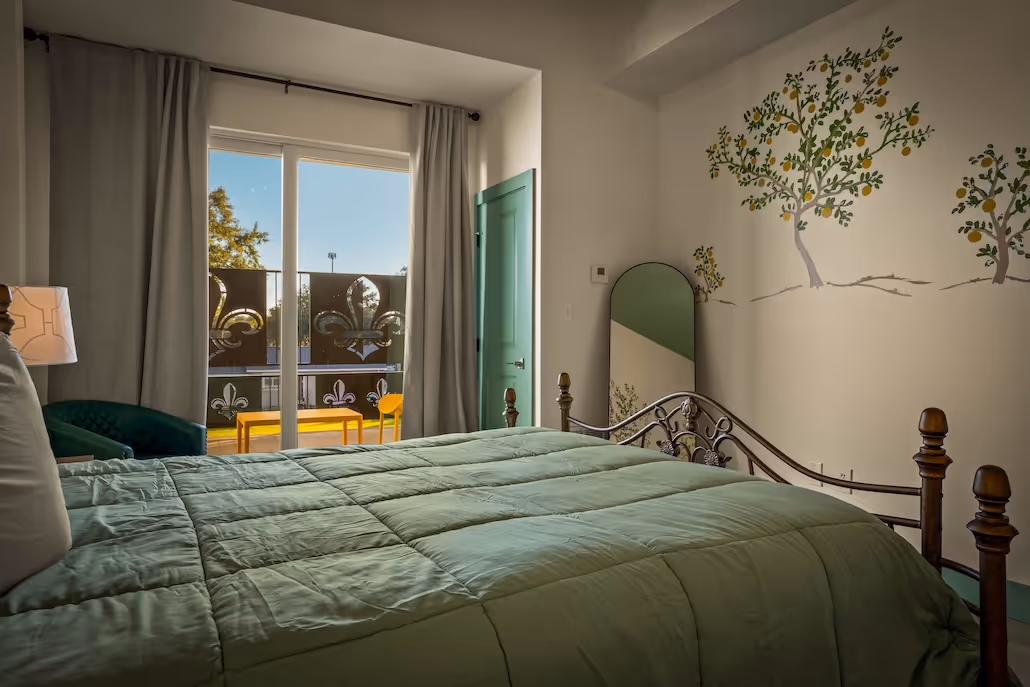 Bedroom with green bedspread, decorative wall mirror, painted tree murals, sliding glass door with fleur-de-lis balcony panels, and teal door.