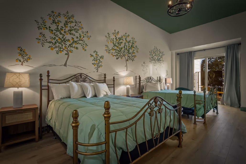 Bedroom with two wrought iron beds with green bedspreads, bedside tables with lamps, and wall paintings of lemon trees.