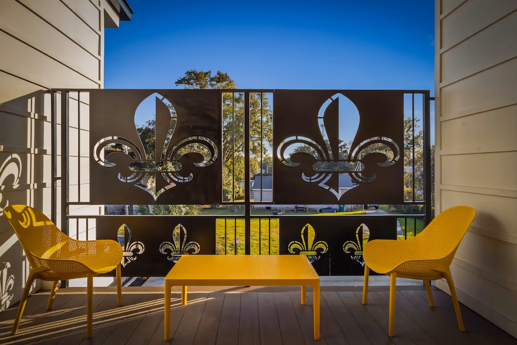 Balcony with yellow chairs and table, metal railing with fleur-de-lis cutouts, and a sunny, clear sky view.