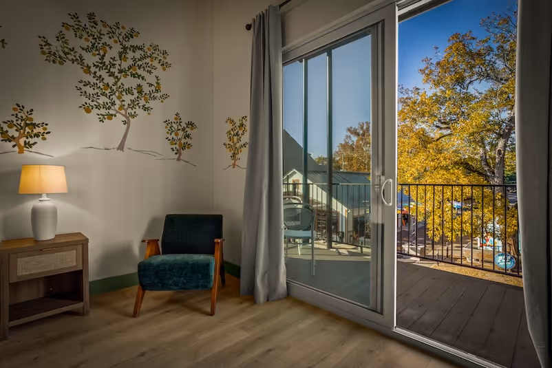Cozy corner with a green upholstered chair, wooden nightstand with a lamp, wall decals of trees, and a sliding glass door leading to a balcony with autumn foliage outside.