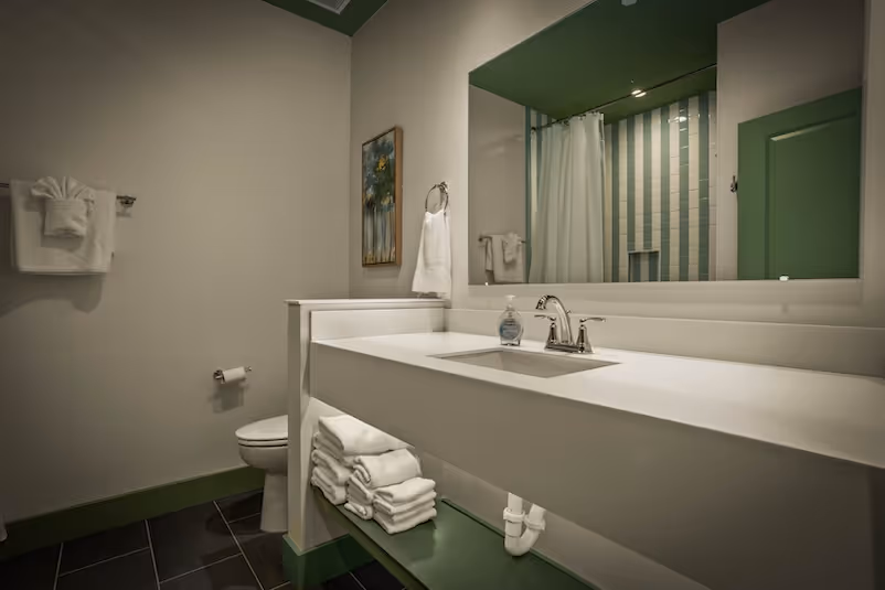 Modern bathroom with white countertop sink, large mirror, green accents, stacked white towels, and a toilet.