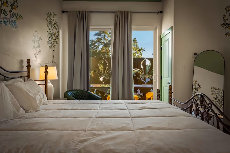 Bedroom with a large white bed, green cushioned chair by a window with gray curtains, floral wall decals, and a tall green-framed mirror.