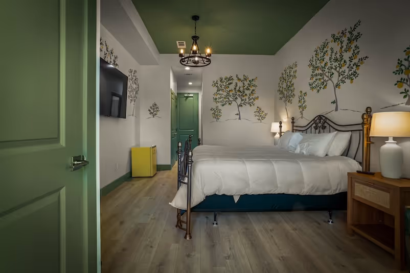 Bedroom with green ceiling, white walls decorated with tree murals, a metal bed with white bedding, a wooden nightstand with lamp, and a yellow mini-fridge under a wall-mounted TV.