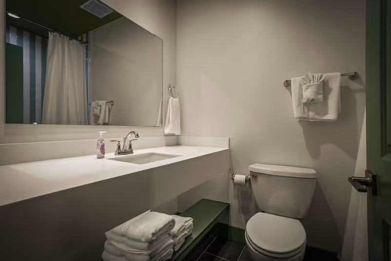 Modern bathroom with white countertop, built-in sink, large mirror, toilet, and neatly folded towels on a green shelf.