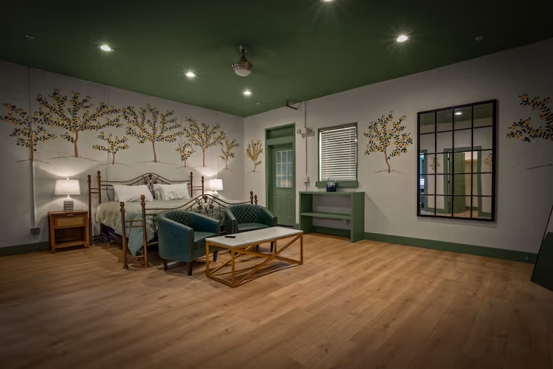 Spacious bedroom with wooden floor, green ceiling, metal bed with white bedding, two green armchairs, a coffee table, bedside table with lamp, wall mirror, and tree mural decorations.