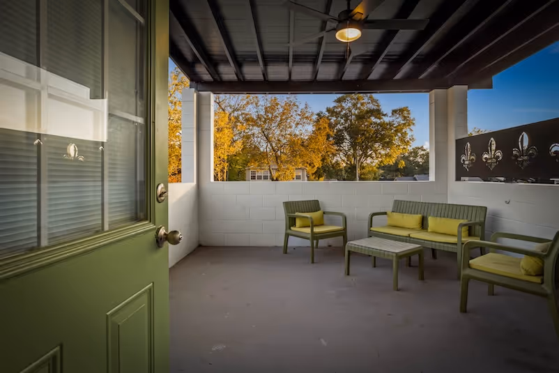 Covered balcony with green cushioned chairs, a small table, and a ceiling fan, overlooking trees with autumn foliage.