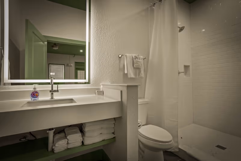Modern bathroom with a white sink, illuminated mirror, stacked towels, toilet, and shower with a white curtain.