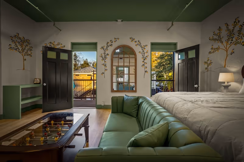 Bedroom with green leather sofa, large bed, foosball table coffee table, open black doors leading to balcony, and wall art of trees around an arched mirror.