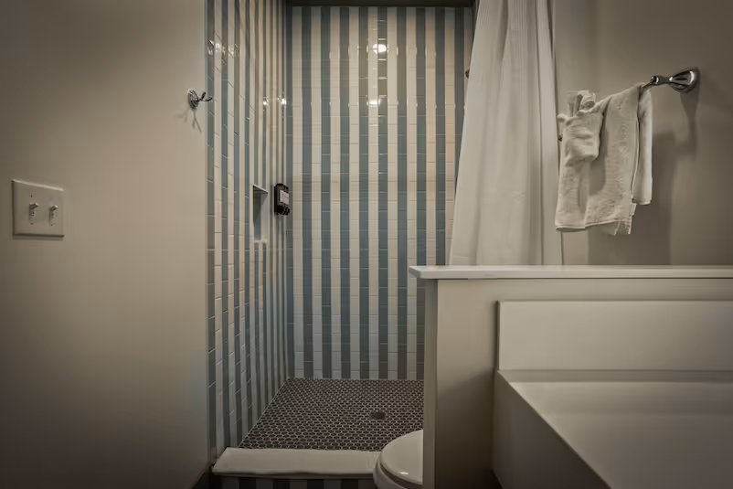 Small bathroom shower area with vertical blue and white striped tiles, white shower curtain, towel rack with folded towels, and partial view of a white sink and toilet.