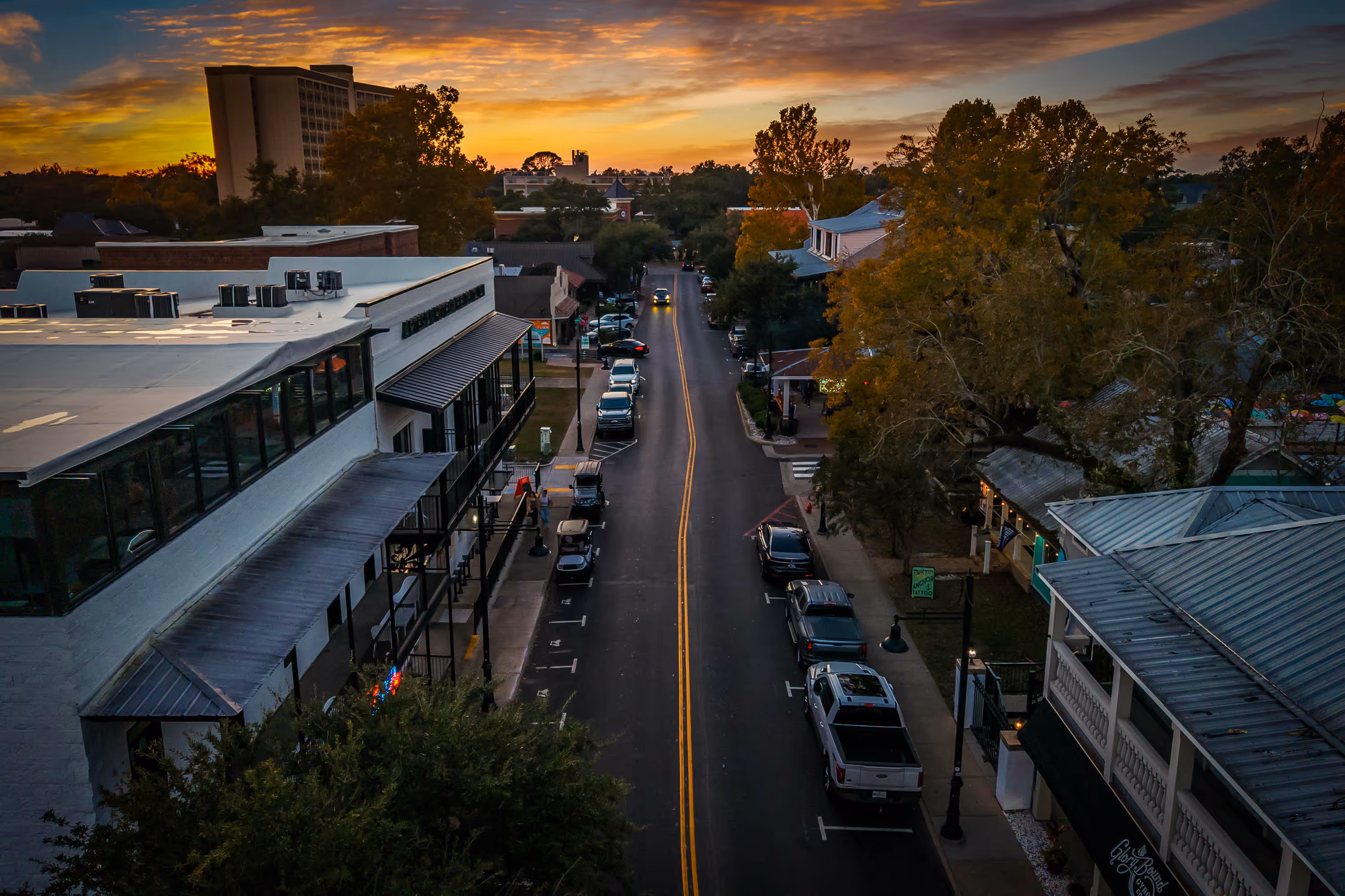 Aerial view of a small town street at sunset with parked cars, buildings, and trees with autumn foliage.