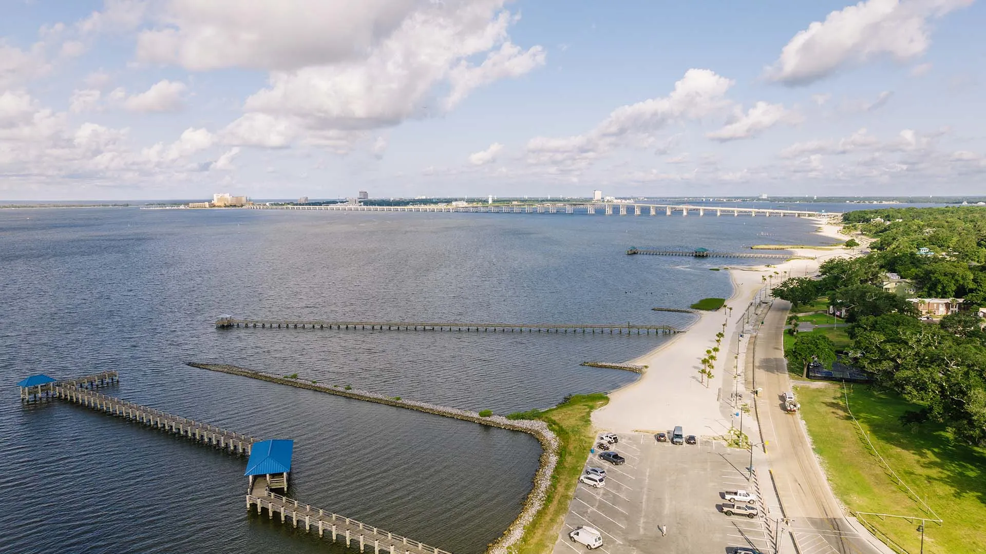 Aerial view of a coastal beach with piers extending into calm water, a parking lot, a road, and green trees under a partly cloudy sky.