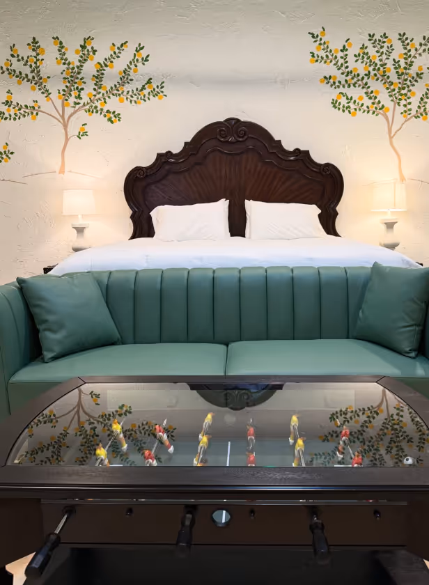 Room with a green sofa, foosball table, large wooden headboard bed, white pillows, and lemon tree wall art with lamps.