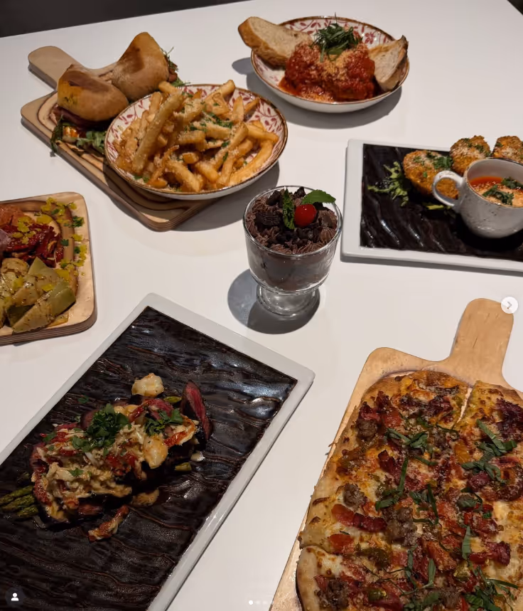 Table with a variety of dishes including a flatbread pizza, crinkle-cut fries, meatballs with tomato sauce and bread, a dessert glass with chocolate and cherry, and plates of assorted appetizers and entrees.