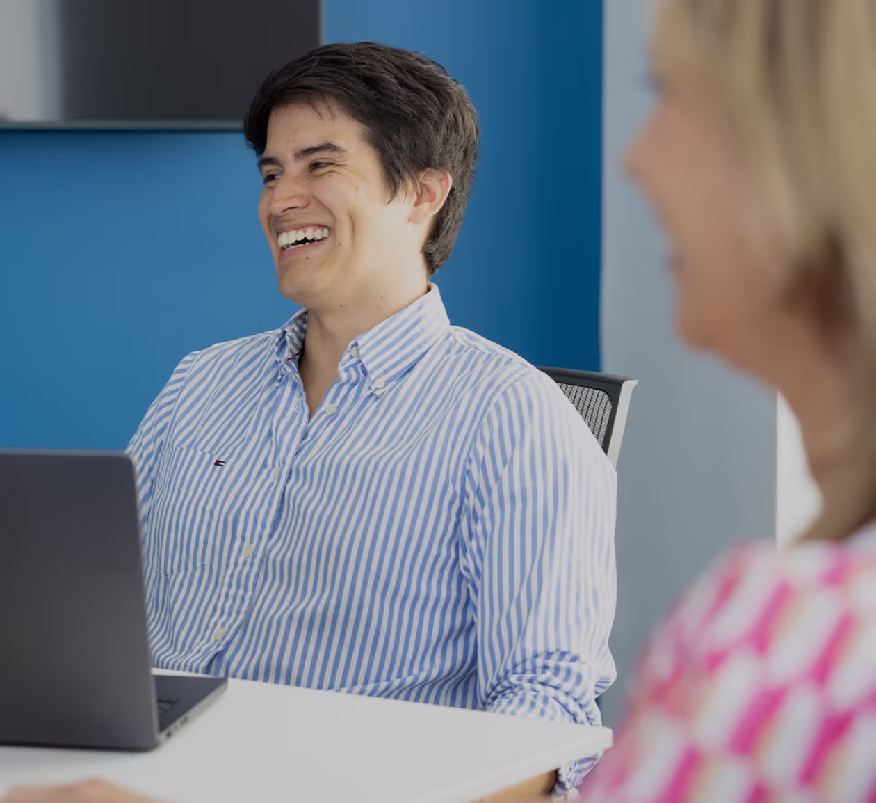 Man in blue and white striped shirt laughing while sitting at a desk with a laptop
