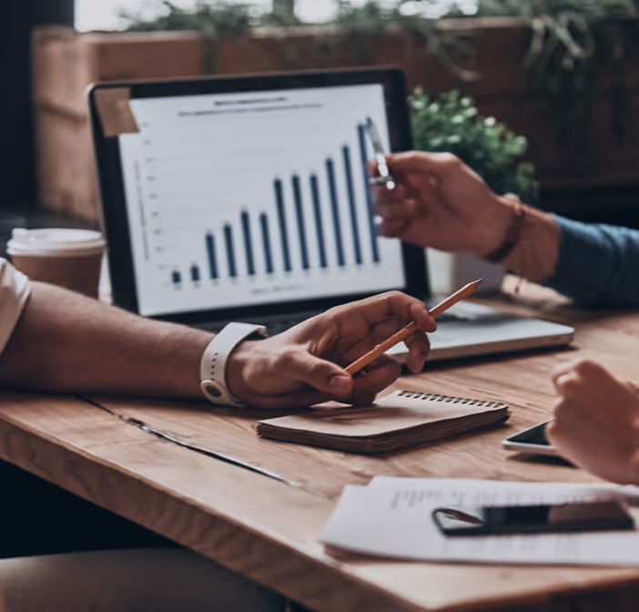 Two people discussing data on a tablet showing a rising bar graph at a wooden table with notebooks and coffee cup.