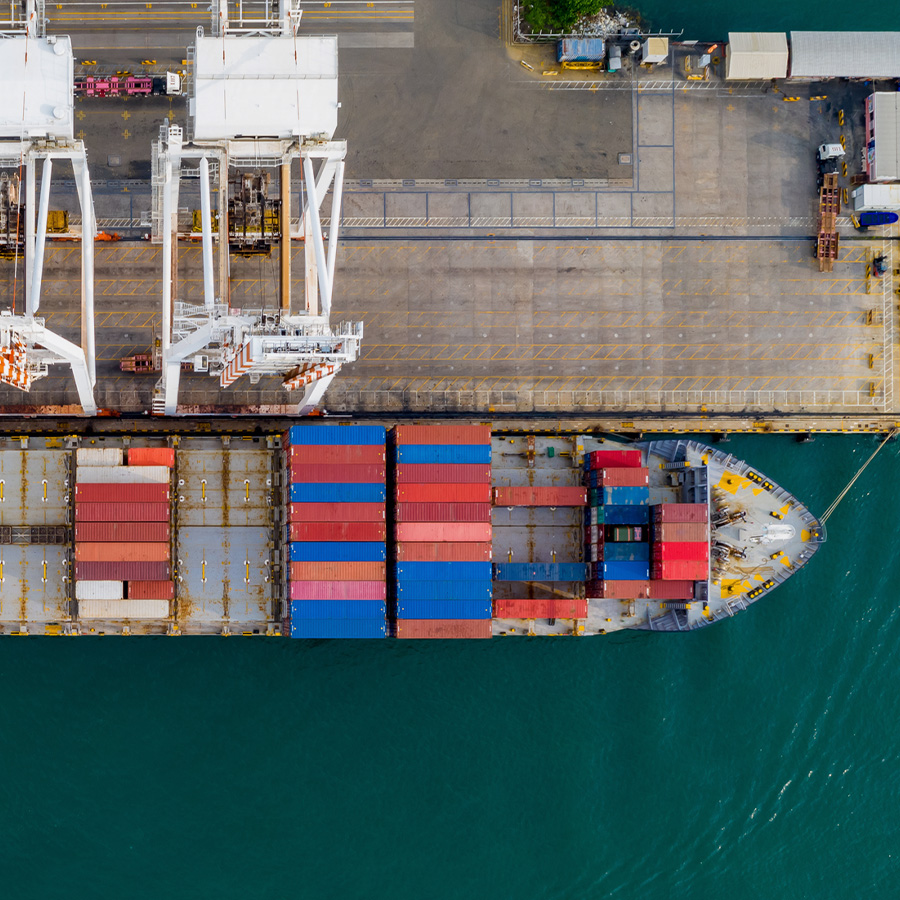 Aerial view of a large container ship loaded with multicolored shipping containers sailing next to a dock with trucks and cranes.