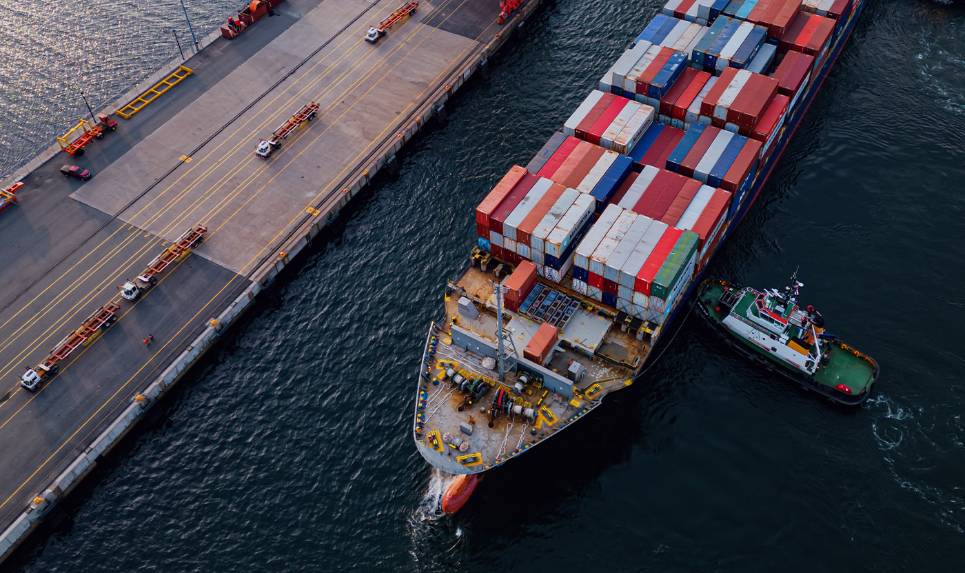 Aerial view of a cargo ship loaded with multicolored shipping containers docked near a pier with small vehicles and a tugboat assisting.