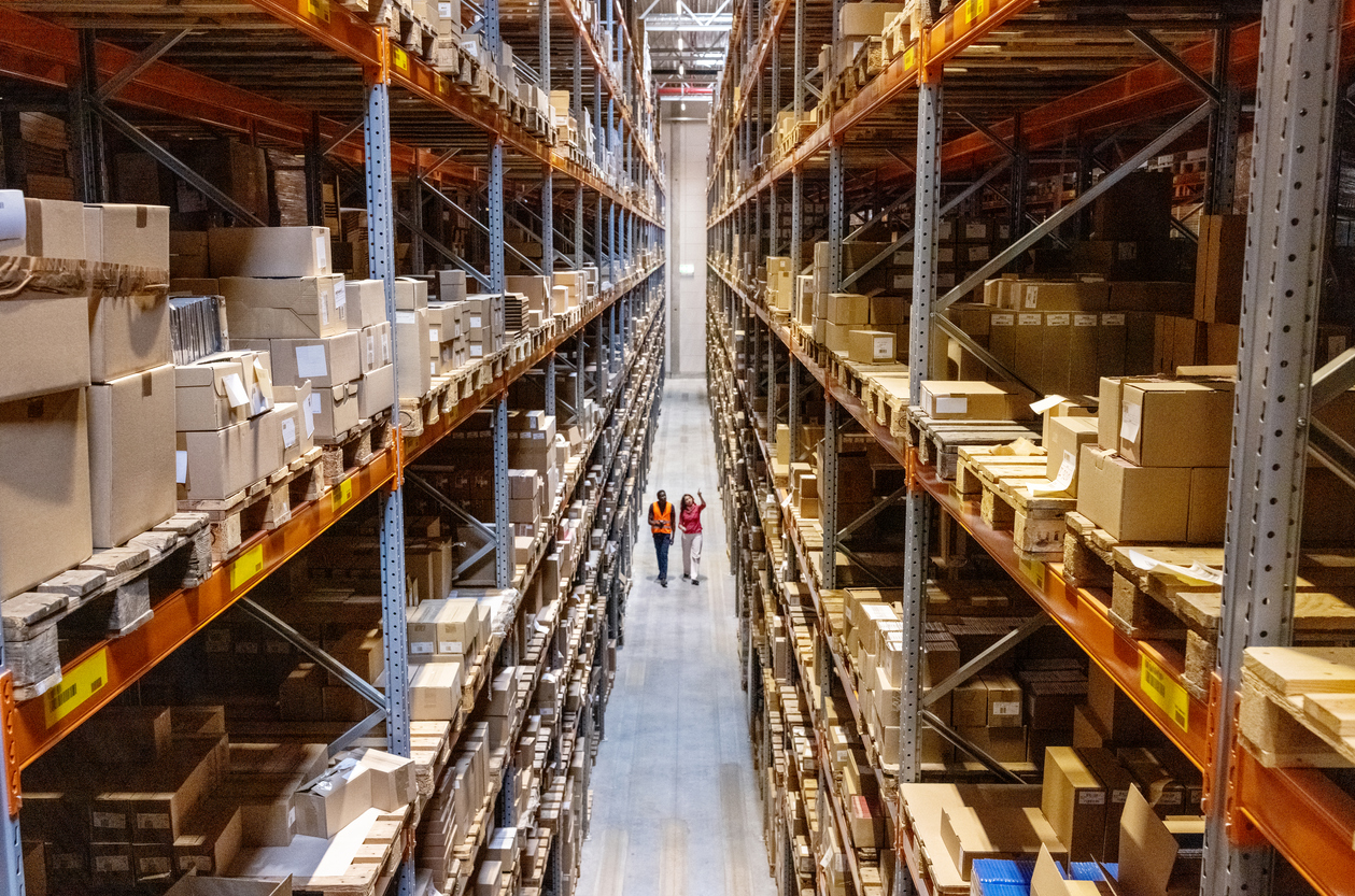 Two workers walking down a narrow aisle between tall warehouse shelves stacked with cardboard boxes.