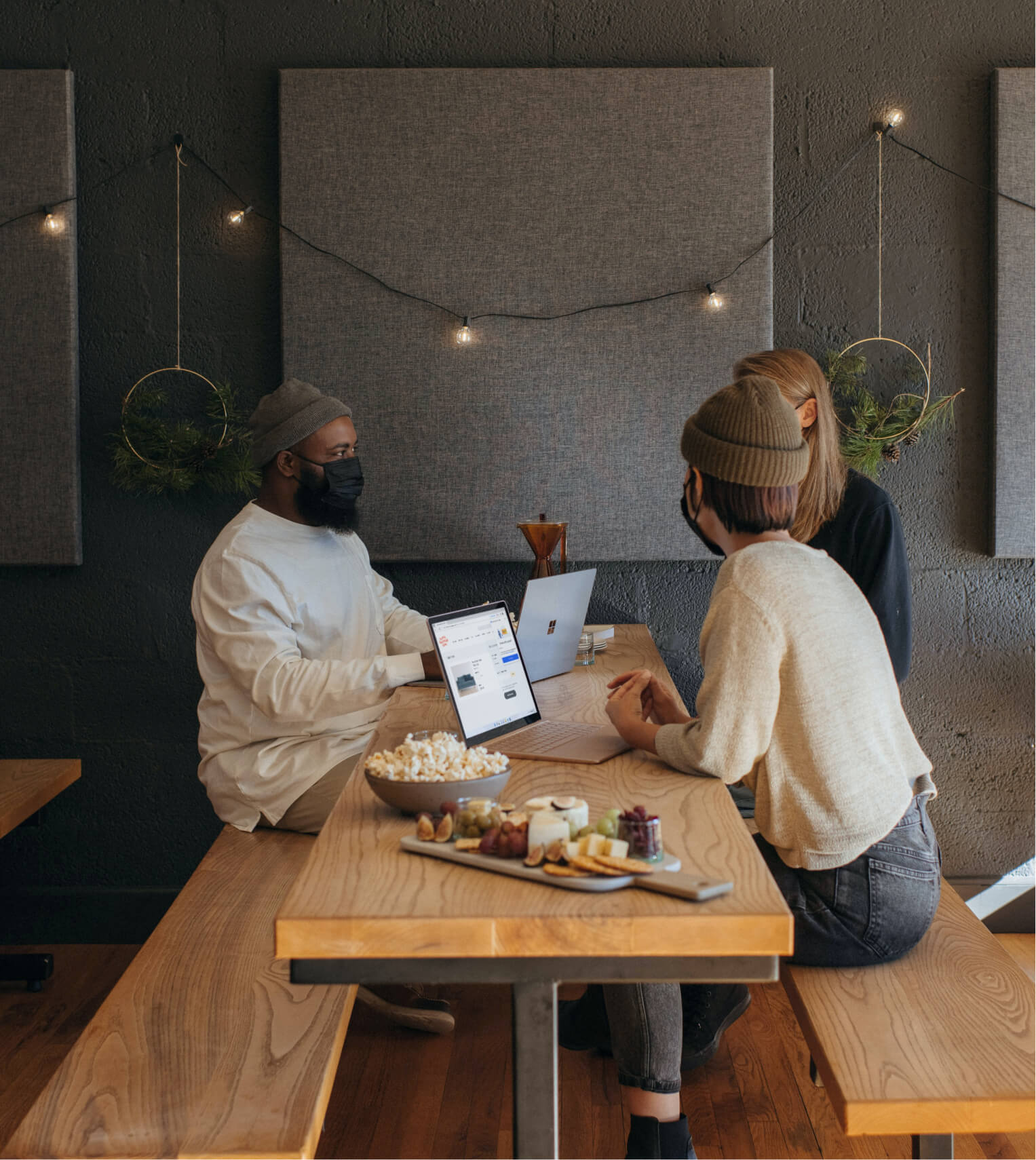 Three people wearing masks sitting at a wooden table with laptops, a bowl of popcorn, and a charcuterie board in a cozy room with gray soundproof panels and string lights on the wall.