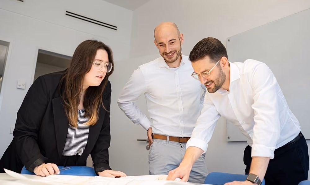 Drei Personen besprechen Pläne auf einem Tisch in einem hellen Büro.