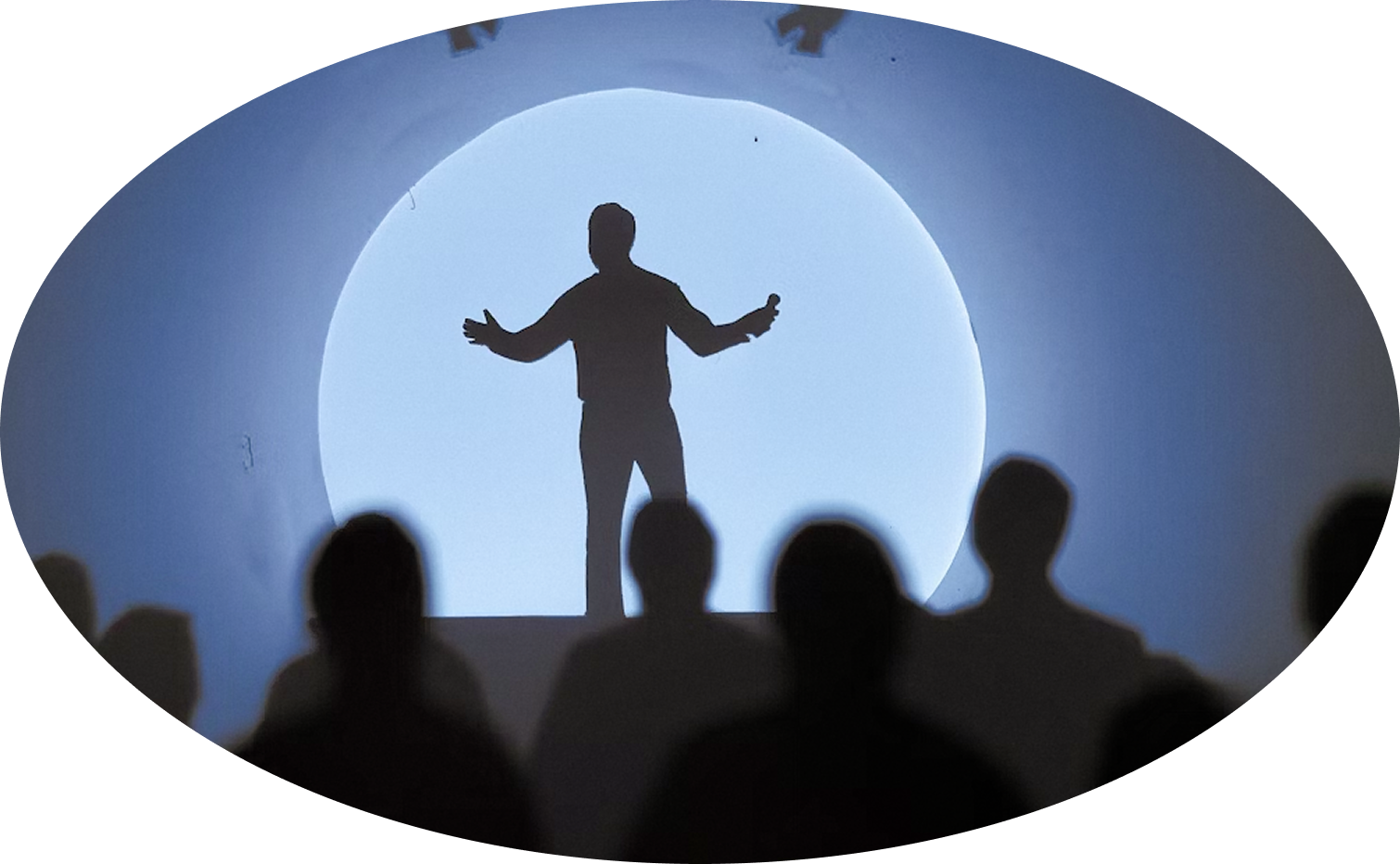 Silhouette of a person speaking with open arms holding a microphone against a large circular light, with blurred audience silhouettes in the foreground.