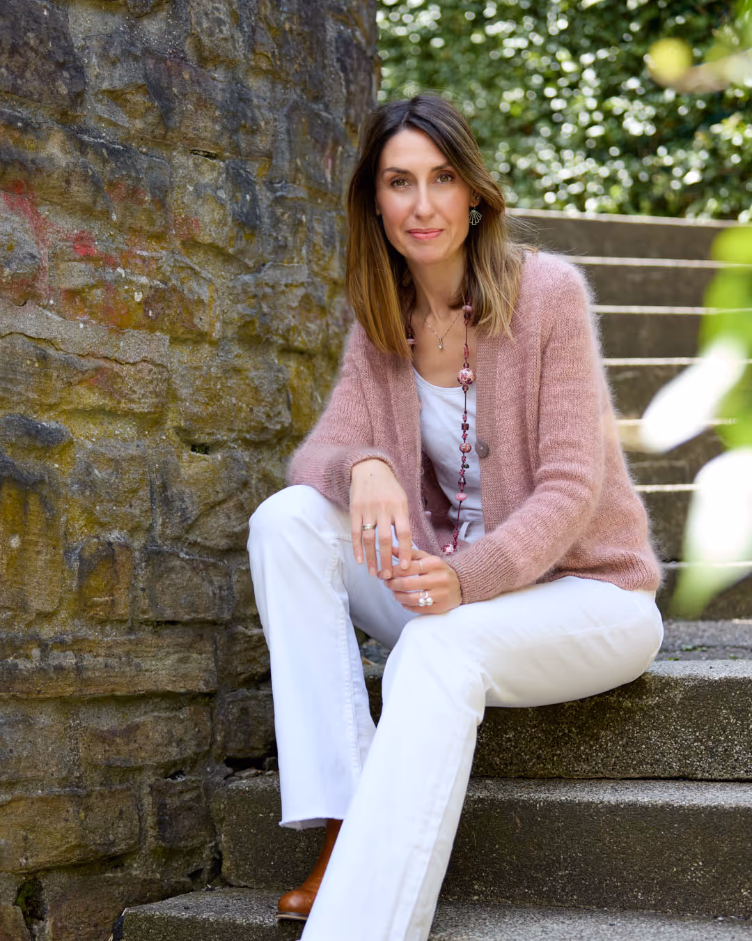 Woman with brown hair wearing a pink cardigan and white pants sitting on stone steps beside a mossy stone wall.