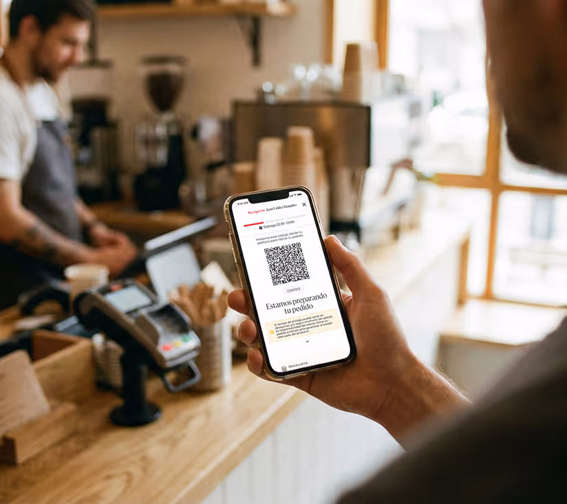 Persona sostiene un celular que muestra una pantalla de preparación de pedido con un código QR en una cafetería.