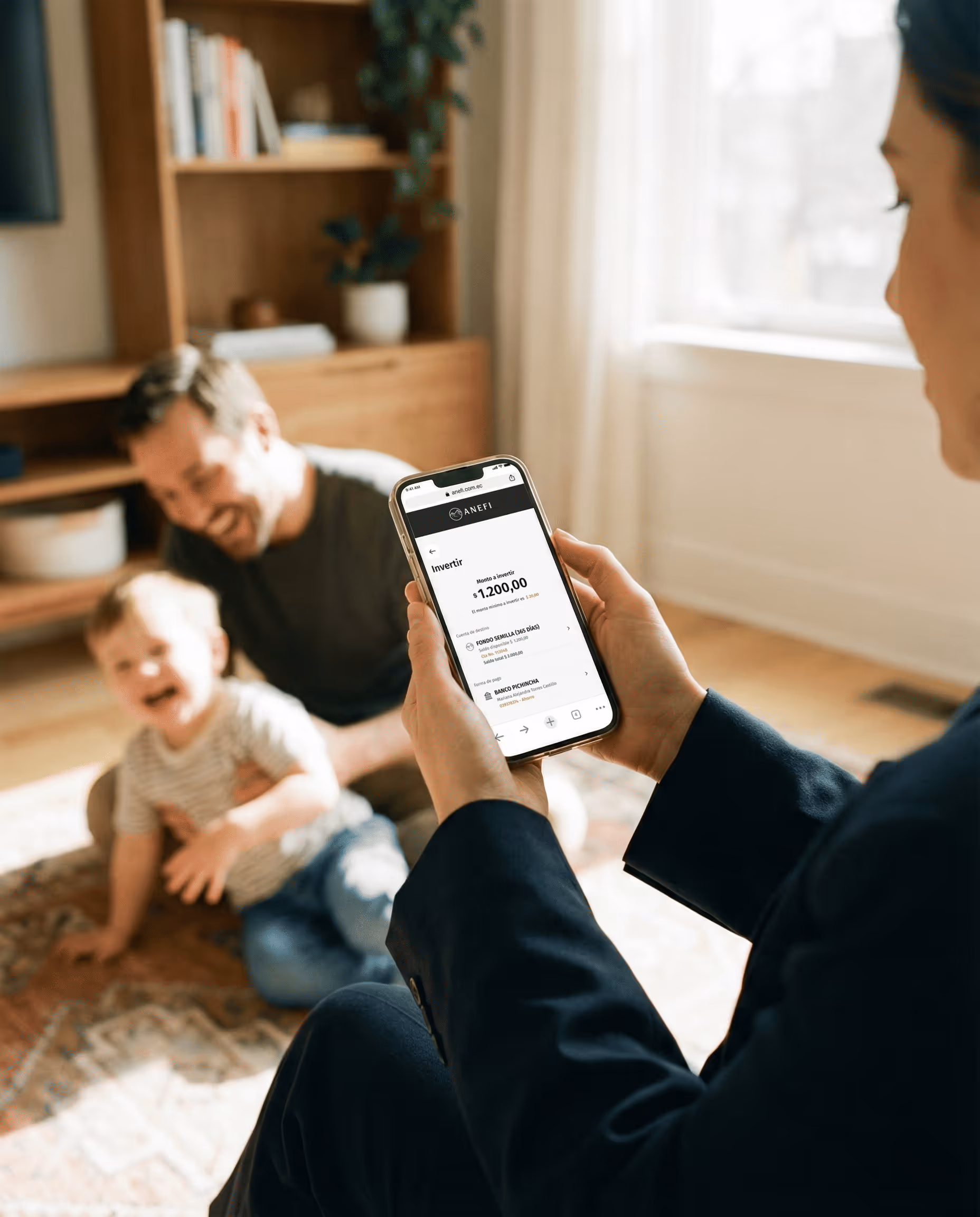 Persona que sostiene un celular que muestra una aplicación de inversión con un monto de $1200, mientras un niño y un adulto juegan borrosos en el fondo.