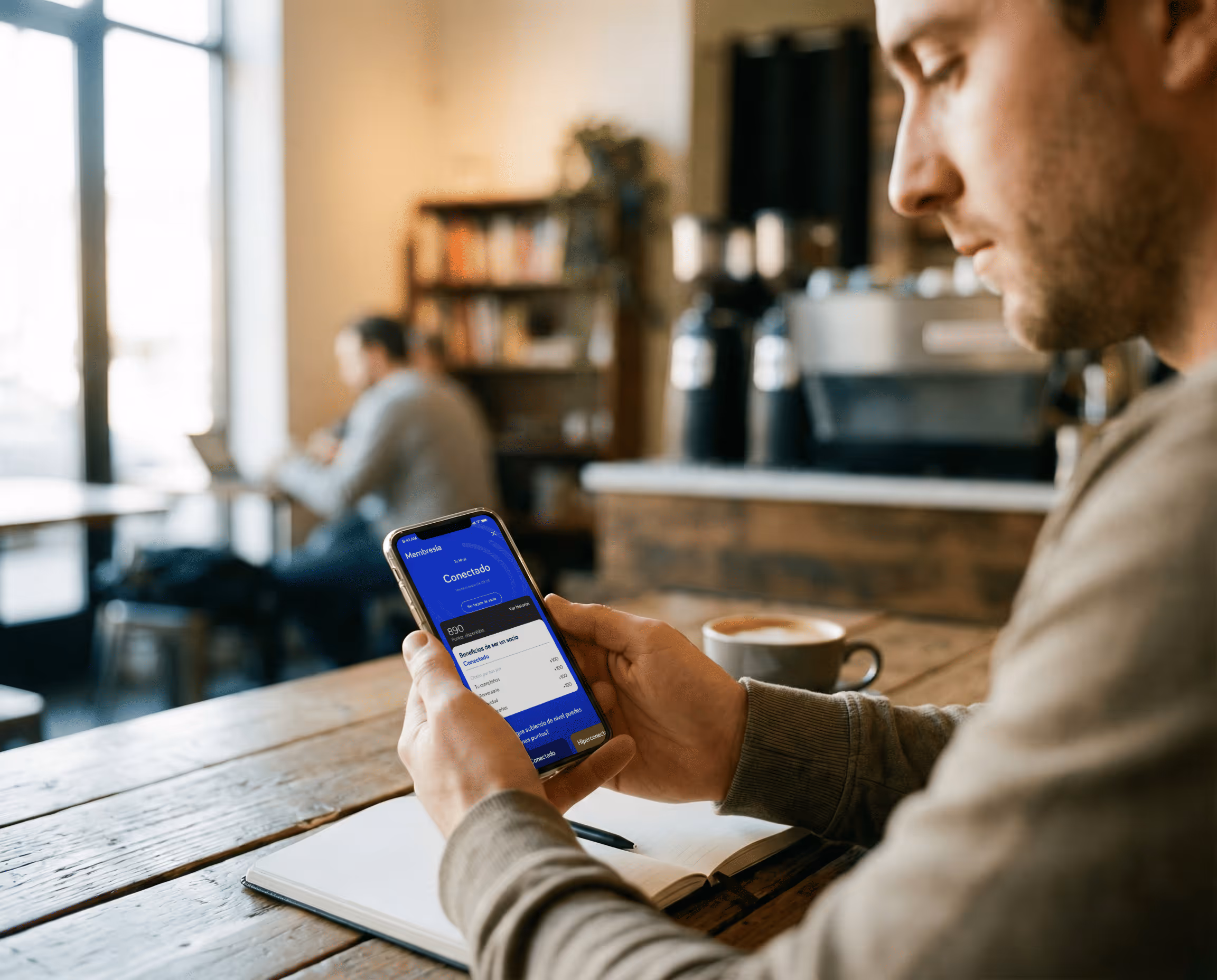 Hombre sentado en un cafe, sostiene su celular mientras observa un programa de puntos de una aplicación de telecomunicaciones.