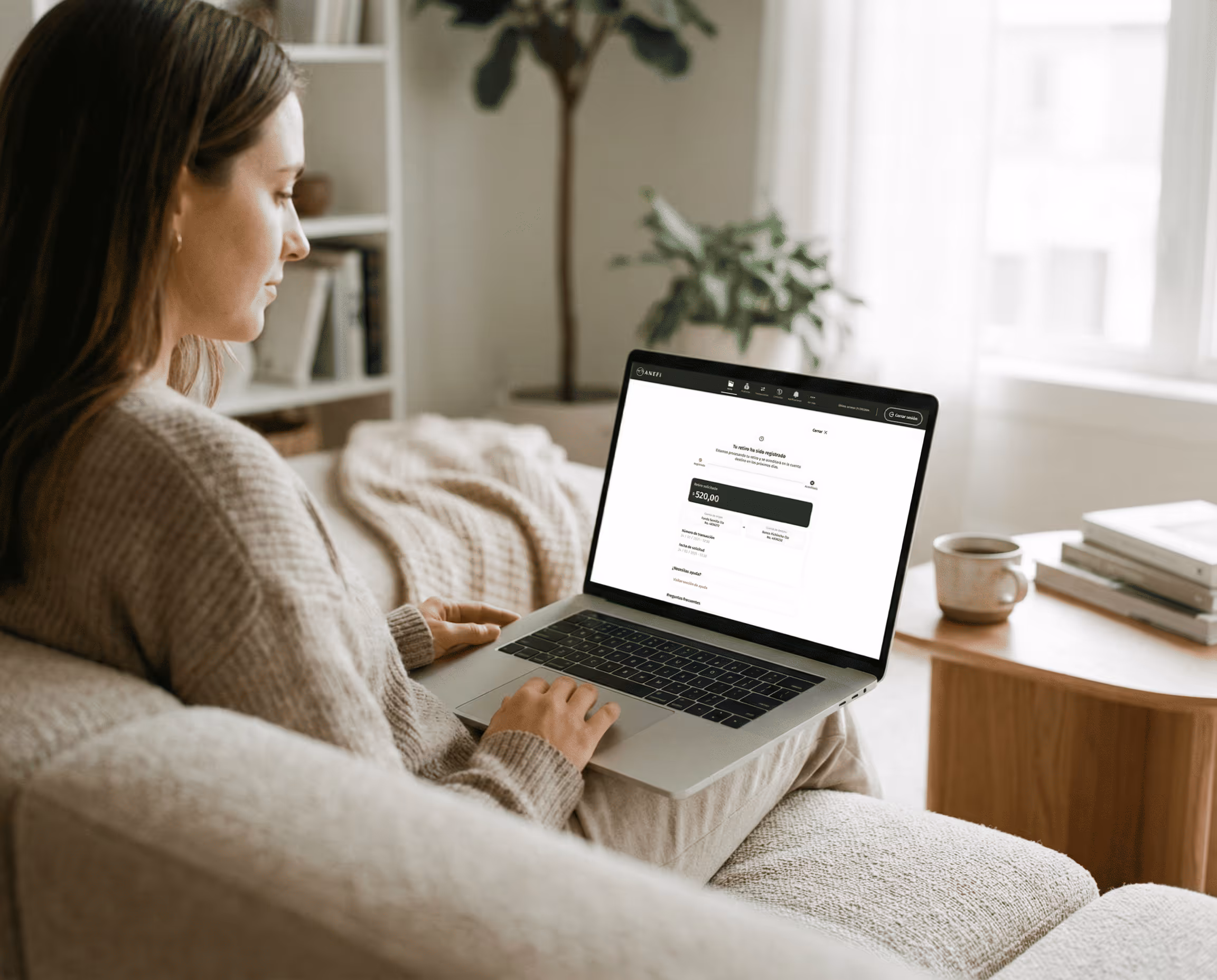 Mujer con un suéter acogedor sentada en un sofá usando una computadora portátil que muestra un tablero financiero con un saldo  de retiro de $ 520,00.