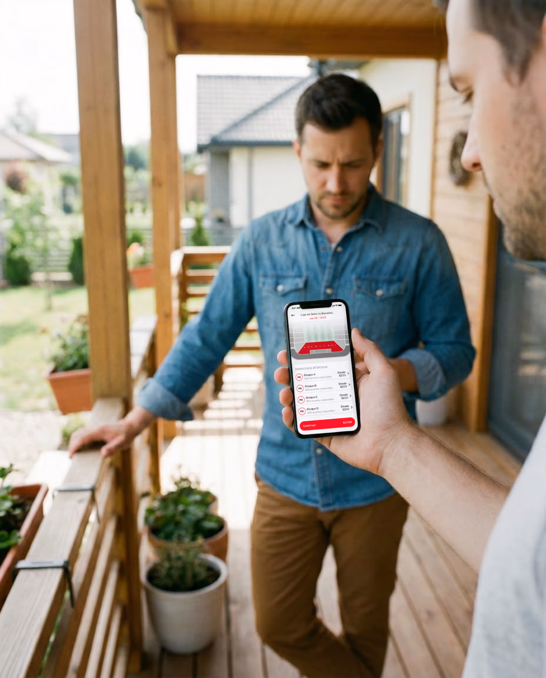 Un hombre sostiene un teléfono que muestra una aplicación de reserva de entradas con opciones de asientos para un evento deportivo, mientras otro hombre aparece borroso en el fondo en un porche de madera.