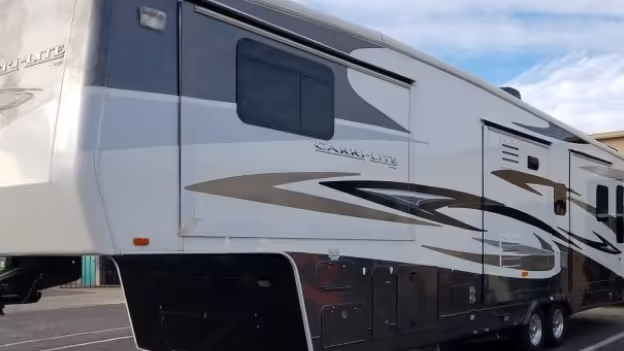 Side view of a large fifth-wheel camper trailer with dark and gray graphics parked on asphalt under a partly cloudy sky.