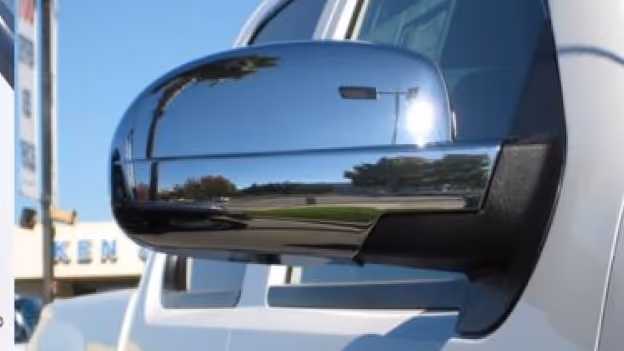 Close-up of a shiny chrome side mirror cap on a white vehicle reflecting nearby trees and a light pole.