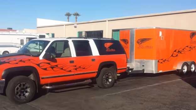 Red and black SUV with flame decals towing a matching red trailer with black logos parked in a sunlit lot.